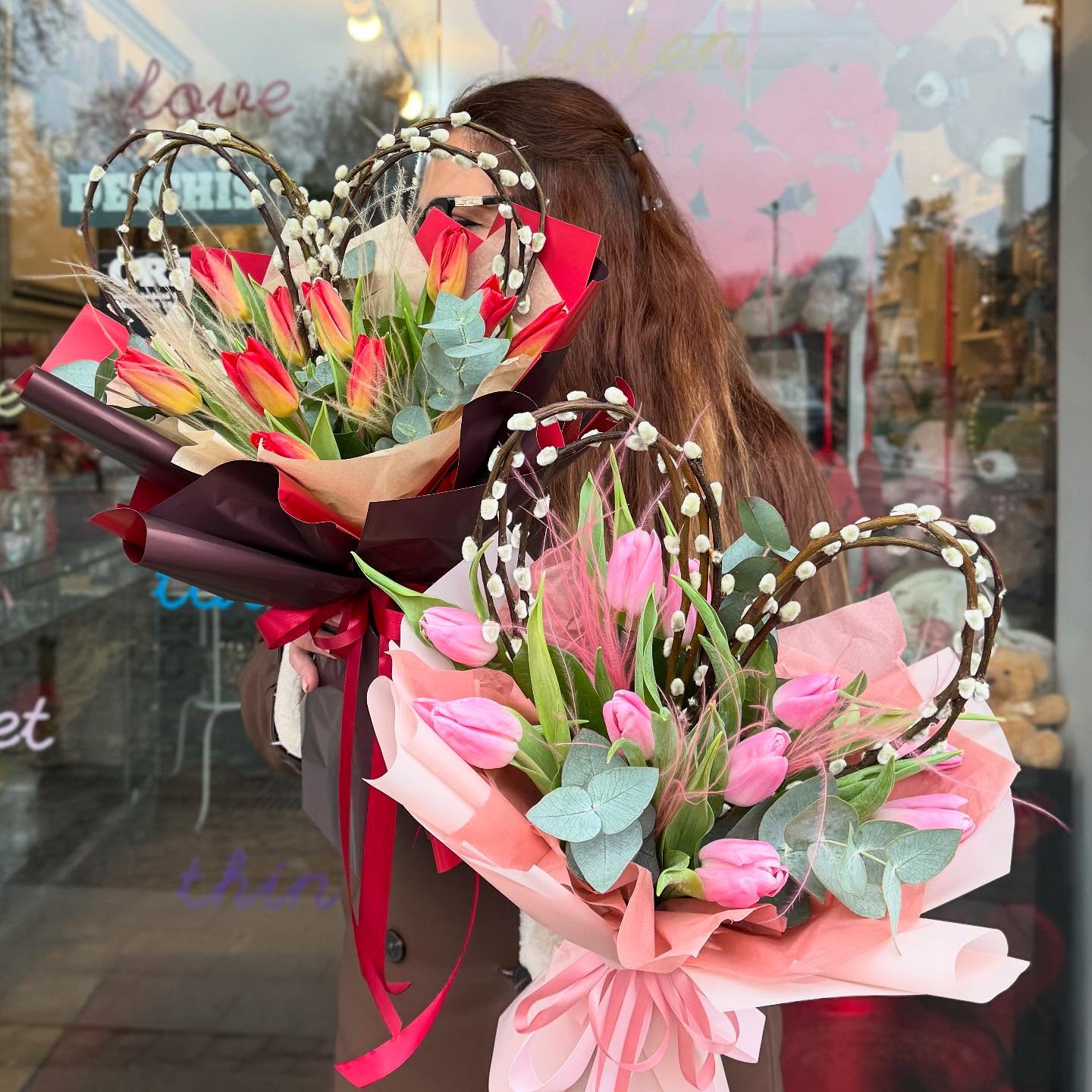 Floristă Floral Studio ținând două buchete de lalele – lalele roșii și lalele roz – cu inimi decorative din răchită, în fața florăriei din Iași
